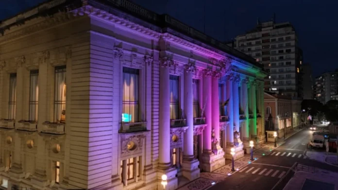 RS inicia semana de conscientização sobre doenças raras Fotografia noturna em ângulo diagonal da fachada lateral e frontal do Palácio Piratini, em Porto Alegre. O prédio histórico de arquitetura clássica está iluminado por projetores de luz nas cores lilás, rosa, azul e verde, cores que simbolizam a campanha do Dia Mundial das Doenças Raras. A iluminação destaca as colunas e detalhes ornamentais da sede do governo, enquanto a rua ao lado aparece com luzes de postes e rastros de veículos ao fundo.
