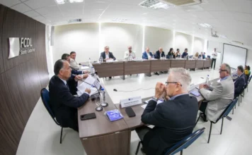Redução de jornada preocupa setor produtivo catarinense Foto em ângulo aberto de uma reunião com diversos líderes empresariais de terno sentados ao redor de uma grande mesa em formato de U, em uma sala de conferência com a identificação "FCDL Santa Catarina" na parede. A imagem ilustra o encontro de membros do Conselho das Federações Empresariais de Santa Catarina (COFEM), mencionado na matéria, debatendo os riscos da redução da jornada de trabalho.