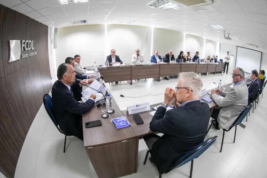 Foto em ângulo aberto de uma reunião com diversos líderes empresariais de terno sentados ao redor de uma grande mesa em formato de U, em uma sala de conferência com a identificação 