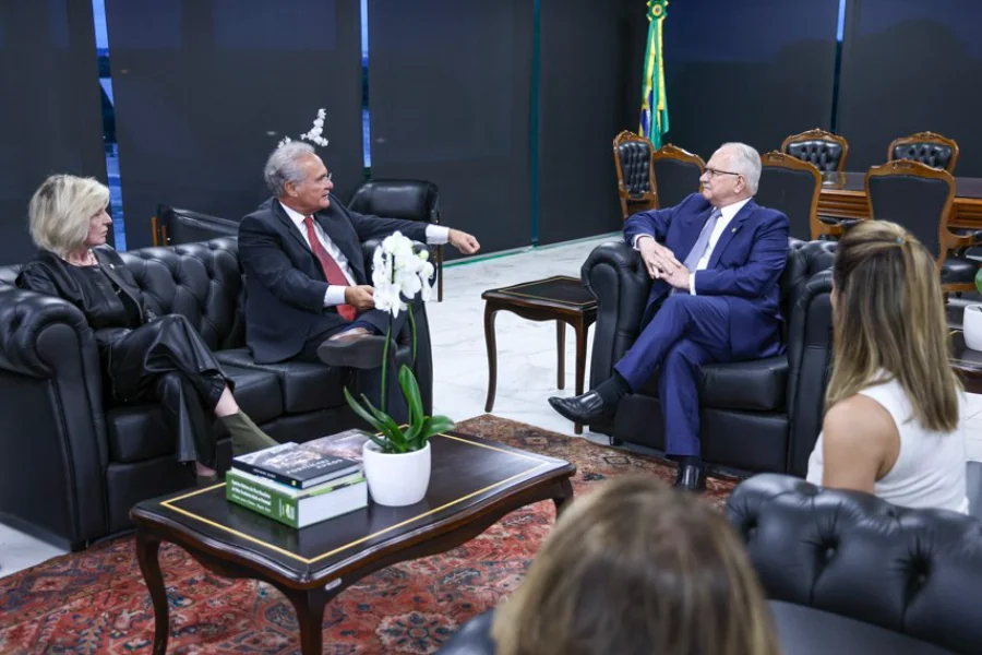 Foto de uma reunião em um gabinete oficial, onde o senador Renan Calheiros (à esquerda, de terno cinza e gravata vermelha) gesticula enquanto conversa com o presidente do STF, ministro Edson Fachin (à direita, de terno azul). Ambos estão sentados em poltronas de couro preto, separados por uma mesa de centro com uma orquídea branca. Outras pessoas, incluindo uma mulher de preto à esquerda e outra de costas em primeiro plano, participam do encontro, que tratou do acesso a investigações sigilosas sobre o Banco Master.