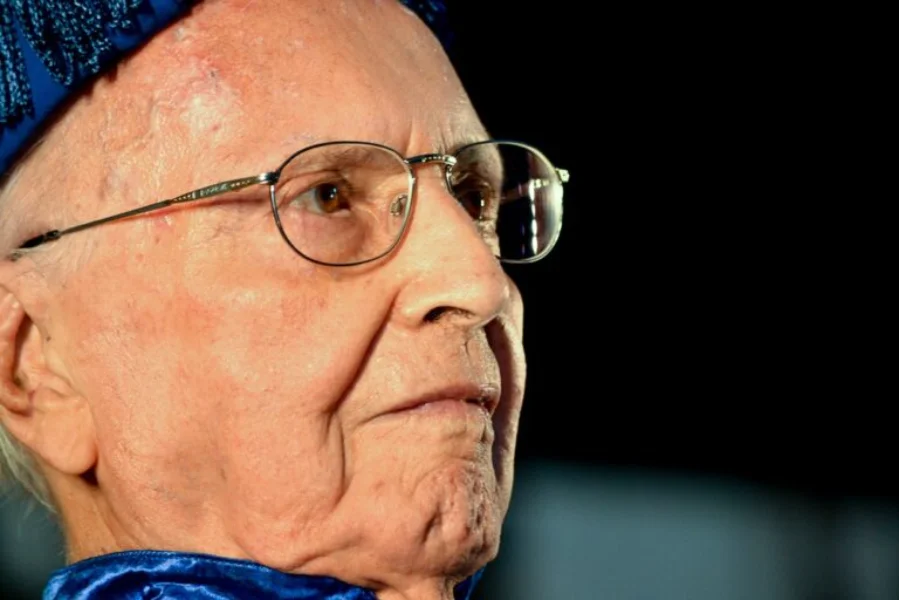 Retrato em close de perfil do ex-prefeito Ruy Hülse, fundador da Unesc, um senhor idoso de óculos, vestindo trajes acadêmicos em tons de azul. Ele possui uma expressão serena e o olhar voltado para o horizonte, simbolizando seu legado centenário.