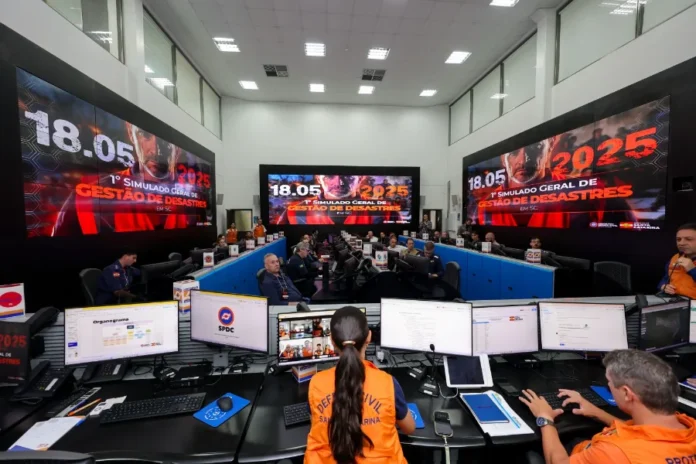 SC abre inscrições para simulado estadual de desastres Centro de operações da Defesa Civil de Santa Catarina, com agentes de coletes laranja trabalhando em estações com múltiplos monitores. Ao fundo, grandes telões exibem a identidade visual do "1º Simulado Geral de Gestão de Desastres em SC - 2025", ilustrando o tipo de estrutura de comando que será mobilizada novamente para o segundo exercício.