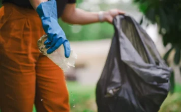 SC confirma 2.097 casos de dengue e alerta para infestação Pessoa utilizando luvas de proteção azuis segura um recipiente plástico pingando água e o descarta em um saco de lixo preto, ilustrando uma ação de limpeza para eliminar focos do mosquito Aedes aegypti.