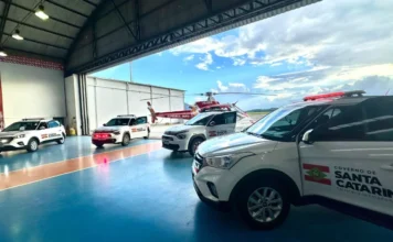 SC entrega novos veículos para reforçar transplantes Vista do interior de um hangar mostrando quatro novos veículos brancos, modelo SUV, com a identidade visual do Governo de Santa Catarina e da Secretaria de Estado da Saúde. Ao fundo, no pátio externo sob um céu nublado, um helicóptero vermelho do SAMU/Corpo de Bombeiros está estacionado, reforçando a integração logística entre o transporte terrestre e o aeromédico para a Central de Transplantes.