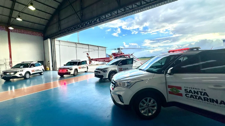 Vista do interior de um hangar mostrando quatro novos veículos brancos, modelo SUV, com a identidade visual do Governo de Santa Catarina e da Secretaria de Estado da Saúde. Ao fundo, no pátio externo sob um céu nublado, um helicóptero vermelho do SAMU/Corpo de Bombeiros está estacionado, reforçando a integração logística entre o transporte terrestre e o aeromédico para a Central de Transplantes.