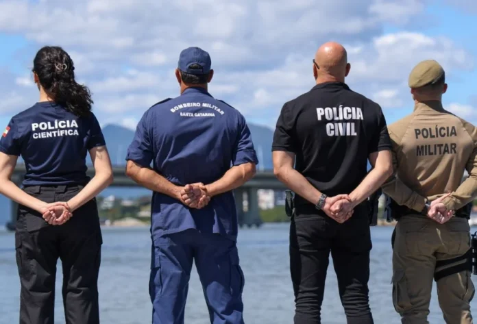 SC inicia 2026 como o estado mais seguro do Brasil Foto tirada pelas costas mostra quatro agentes de segurança pública de Santa Catarina em pé, lado a lado, olhando para uma paisagem de água e pontes. Da esquerda para a direita, uma mulher da Polícia Científica, um homem do Corpo de Bombeiros Militar, um homem da Polícia Civil e um homem da Polícia Militar. A imagem representa a integração das forças policiais que contribui para que o estado seja o mais seguro do Brasil.