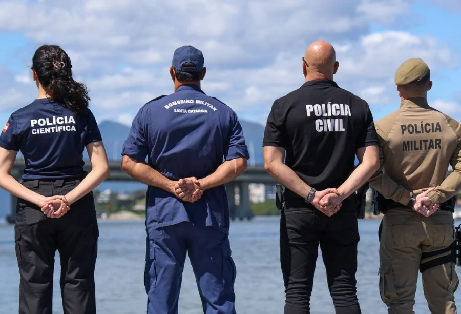 Foto tirada pelas costas mostra quatro agentes de segurança pública de Santa Catarina em pé, lado a lado, olhando para uma paisagem de água e pontes. Da esquerda para a direita, uma mulher da Polícia Científica, um homem do Corpo de Bombeiros Militar, um homem da Polícia Civil e um homem da Polícia Militar. A imagem representa a integração das forças policiais que contribui para que o estado seja o mais seguro do Brasil.