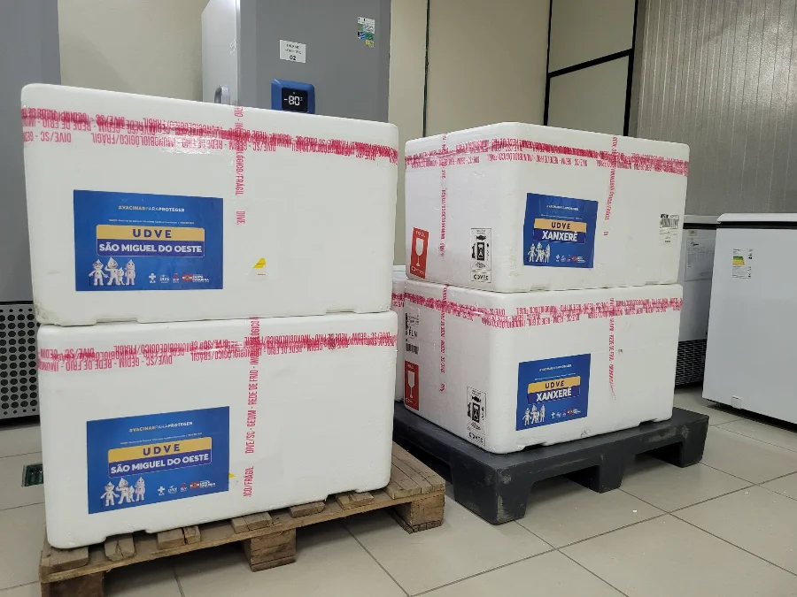 Quatro caixas térmicas de isopor brancas, lacradas com fitas de segurança vermelhas, estão empilhadas sobre paletes de madeira e plástico em um ambiente de armazenamento climatizado. As caixas possuem etiquetas azuis de identificação da Secretaria de Estado da Saúde de Santa Catarina, com os destinos destacados: 