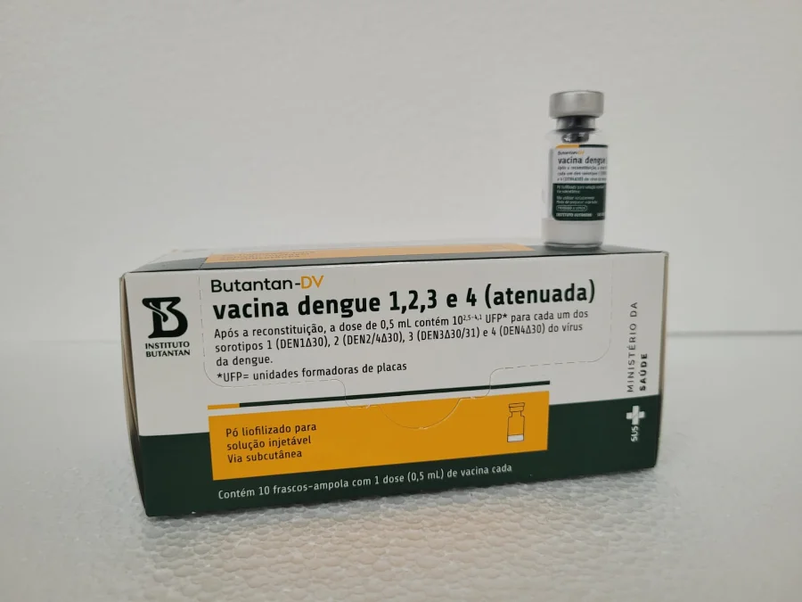 A imagem apresenta, em destaque central, uma caixa de cor branca e laranja do imunizante Butantan-DV, a vacina contra a dengue produzida pelo Instituto Butantan. Na embalagem, lê-se o texto 