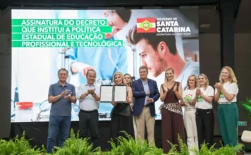 SC lança política de ensino técnico com 50 mil novas vagas Fotografia em plano médio registra o momento da assinatura do decreto que institui a Política Estadual de Educação Profissional e Tecnológica em Santa Catarina. No centro, uma autoridade feminina segura o documento assinado, ladeada pelo governador Jorginho Mello e outros representantes do governo e da educação, que aplaudem o ato. Ao fundo, um grande telão exibe o título do evento em letras brancas sobre faixas verdes, junto ao brasão do Governo do Estado. O palco está decorado com folhagens verdes na parte inferior.