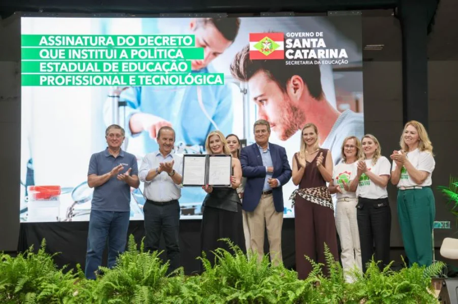 Fotografia em plano médio registra o momento da assinatura do decreto que institui a Política Estadual de Educação Profissional e Tecnológica em Santa Catarina. No centro, uma autoridade feminina segura o documento assinado, ladeada pelo governador Jorginho Mello e outros representantes do governo e da educação, que aplaudem o ato. Ao fundo, um grande telão exibe o título do evento em letras brancas sobre faixas verdes, junto ao brasão do Governo do Estado. O palco está decorado com folhagens verdes na parte inferior.