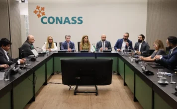 SC lidera transformação digital do SUS em seminário nacional Fotografia em plano médio de uma sala de reuniões no Conass, em Brasília. Ao centro, uma mesa em formato de "U" acomoda cerca de dez gestores e autoridades em trajes formais. Ao fundo, na parede clara, destaca-se o logotipo oficial do Conass em verde e laranja. À frente da mesa, há um grande monitor de televisão voltado para os participantes. O grupo está em meio a uma discussão sobre a transformação digital no SUS.