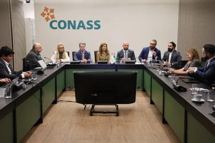 SC lidera transformação digital do SUS em seminário nacional Fotografia em plano médio de uma sala de reuniões no Conass, em Brasília. Ao centro, uma mesa em formato de "U" acomoda cerca de dez gestores e autoridades em trajes formais. Ao fundo, na parede clara, destaca-se o logotipo oficial do Conass em verde e laranja. À frente da mesa, há um grande monitor de televisão voltado para os participantes. O grupo está em meio a uma discussão sobre a transformação digital no SUS.
