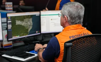 SC mobiliza 98% das cidades para simulado de desastres Homem de perfil, com cabelos grisalhos e óculos, veste um colete laranja oficial da Defesa Civil. Ele está sentado em uma sala de operação diante de dois monitores. A tela da esquerda exibe o mapa de Santa Catarina dividido por municípios, sinalizando o monitoramento geográfico para o simulado estadual. O ambiente sugere uma central de gerenciamento de crises e planejamento logístico.
