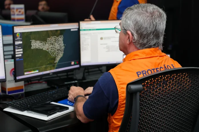 SC mobiliza 98% das cidades para simulado de desastres Homem de perfil, com cabelos grisalhos e óculos, veste um colete laranja oficial da Defesa Civil. Ele está sentado em uma sala de operação diante de dois monitores. A tela da esquerda exibe o mapa de Santa Catarina dividido por municípios, sinalizando o monitoramento geográfico para o simulado estadual. O ambiente sugere uma central de gerenciamento de crises e planejamento logístico.