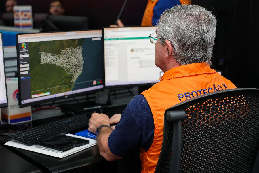 Homem de perfil, com cabelos grisalhos e óculos, veste um colete laranja oficial da Defesa Civil. Ele está sentado em uma sala de operação diante de dois monitores. A tela da esquerda exibe o mapa de Santa Catarina dividido por municípios, sinalizando o monitoramento geográfico para o simulado estadual. O ambiente sugere uma central de gerenciamento de crises e planejamento logístico.