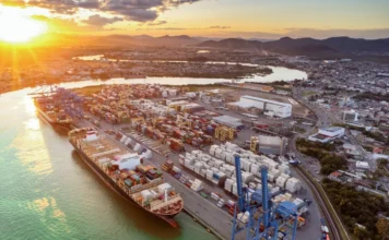 SC movimenta 19,1% dos contêineres do Brasil em 2025 Vista aérea panorâmica de um grande terminal portuário movimentado em Santa Catarina durante o pôr do sol. Um imenso navio porta-contêineres está atracado no cais, sendo carregado por guindastes azuis, enquanto milhares de contêineres coloridos estão empilhados no pátio logístico, ilustrando a alta capacidade de movimentação de cargas do estado descrita na matéria.