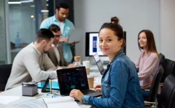 SC oferece curso gratuito de IA para desenvolvedores A imagem apresenta um grupo de jovens profissionais em um ambiente de escritório moderno e iluminado, utilizando computadores portáteis em uma mesa de trabalho compartilhada. Em destaque, uma mulher de jaqueta jeans sorri para a câmera, representando o público-alvo de desenvolvedores de software em Santa Catarina interessados em integrar a inteligência artificial ao seu fluxo de trabalho por meio do curso gratuito oferecido pelo programa SCTEC.