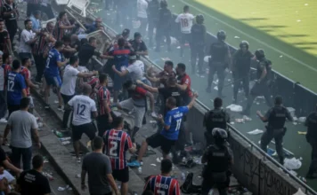 SC sanciona leis contra racismo e violência nos estádios Cena de confusão generalizada nas arquibancadas de um estádio de futebol, mostrando um confronto entre dezenas de torcedores e a intervenção de policiais militares com equipamentos de tropa de choque. Há fumaça visível no ar, ilustrando o tipo de violência que a nova legislação de Santa Catarina busca combater com multas individuais.