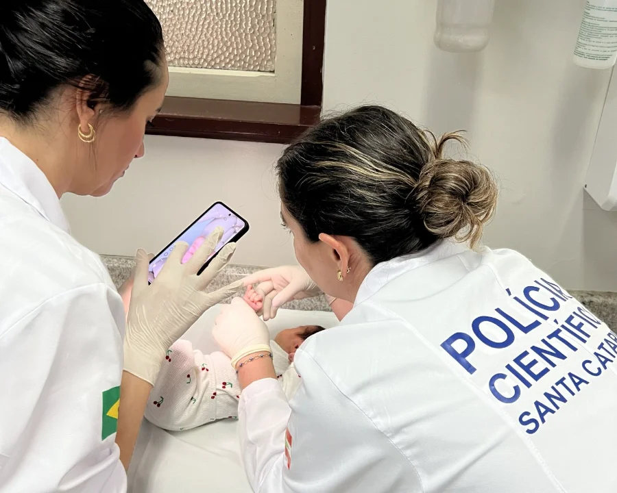 Duas profissionais da Polícia Científica de Santa Catarina, vestindo jalecos brancos e luvas, realizam o procedimento de coleta biométrica e registro fotográfico de um recém-nascido em uma maternidade. Uma das servidoras utiliza um smartphone para capturar a imagem do bebê, enquanto a outra auxilia no posicionamento da criança sobre o trocador.