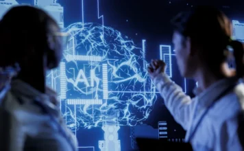 SENAI/SC encerra ciclo sobre IA nesta quinta-feira A imagem apresenta dois profissionais em silhueta observando uma tela digital em tons de azul. Ao centro, destaca-se a representação de um cérebro humano composto por conexões neurais tecnológicas e circuitos, com a sigla "AI" (Inteligência Artificial) em evidência no meio do órgão, simbolizando a integração entre tecnologia e intelecto humano abordada pelo SENAI/SC.