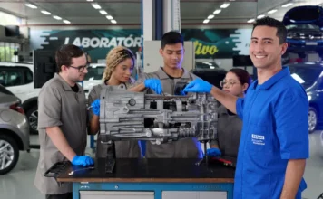 SENAI/SC registra 155 mil matrículas em cursos em 2025 Estudantes do SENAI/SC realizam uma atividade prática em uma oficina de manutenção automotiva. No centro da imagem, quatro jovens com uniformes cinza e luvas azuis analisam uma transmissão mecânica sobre uma bancada de metal. Em primeiro plano, à direita, um estudante com o uniforme azul do SENAI sorri para a câmera. Ao fundo, é possível ver um ambiente de laboratório com carros e equipamentos industriais.