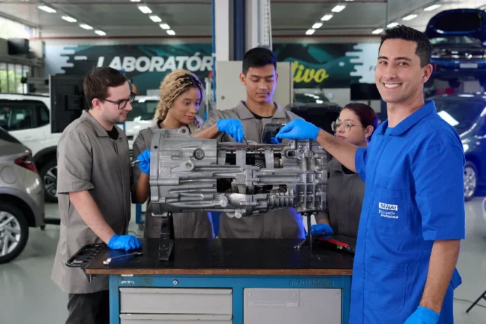 SENAI SC registra 155 mil matrículas em cursos em 2025 Estudantes do SENAI/SC realizam uma atividade prática em uma oficina de manutenção automotiva. No centro da imagem, quatro jovens com uniformes cinza e luvas azuis analisam uma transmissão mecânica sobre uma bancada de metal. Em primeiro plano, à direita, um estudante com o uniforme azul do SENAI sorri para a câmera. Ao fundo, é possível ver um ambiente de laboratório com carros e equipamentos industriais.