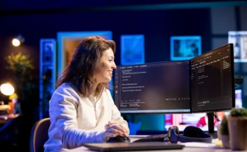 SENAI promove encontro online sobre Análise de Dados Uma profissional sorrindo enquanto trabalha em dois monitores com código, ilustrando o ambiente de tecnologia e análise de dados, tema do encontro online promovido pelo SENAI/SC.