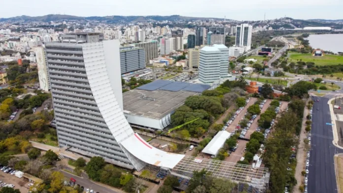 SES alerta para fim de adesão ao Geração Consciente 2026 no RS Vista aérea do Centro Administrativo Fernando Ferrari (CAFF), em Porto Alegre, onde está sediada a Secretaria da Saúde do Rio Grande do Sul. O prédio, com sua icônica fachada branca e curva, aparece em destaque sob um céu claro, cercado por vegetação e outras edificações governamentais. A imagem simboliza a gestão estadual responsável pela coordenação do programa Geração Consciente.
