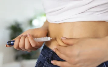 SES alerta para riscos de “canetas” emagrecedoras em SC Close-up da região abdominal de uma mulher vestindo camiseta branca e jeans, enquanto ela aplica uma injeção subcutânea na barriga utilizando um dispositivo semelhante a uma caneta aplicadora. A imagem ilustra o método de uso de medicamentos como a tirzepatida, cujo consumo indiscriminado e sem receita médica é alvo de alerta da Vigilância Sanitária de Santa Catarina devido aos riscos à saúde.