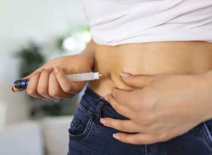 SES alerta para riscos de canetas emagrecedoras em SC Close-up da região abdominal de uma mulher vestindo camiseta branca e jeans, enquanto ela aplica uma injeção subcutânea na barriga utilizando um dispositivo semelhante a uma caneta aplicadora. A imagem ilustra o método de uso de medicamentos como a tirzepatida, cujo consumo indiscriminado e sem receita médica é alvo de alerta da Vigilância Sanitária de Santa Catarina devido aos riscos à saúde.