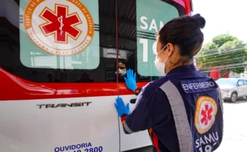 SES lança curso EAD de emergências psiquiátricas em SC Enfermeira do SAMU, vestindo uniforme azul com identificação e máscara, observa o interior de uma ambulância branca e vermelha. A imagem representa os profissionais de urgência que são o público-alvo do novo curso de Emergências Psiquiátricas da SES.