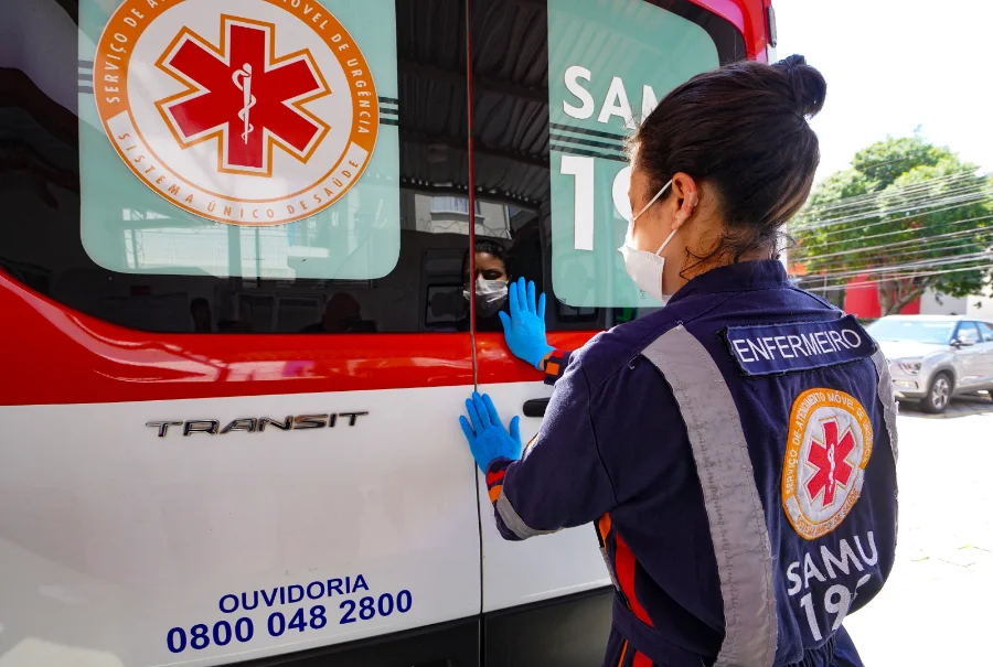 SES lança curso EAD de emergências psiquiátricas em SC Enfermeira do SAMU, vestindo uniforme azul com identificação e máscara, observa o interior de uma ambulância branca e vermelha. A imagem representa os profissionais de urgência que são o público-alvo do novo curso de Emergências Psiquiátricas da SES.