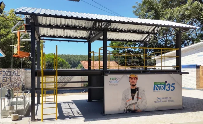 SESI SC registra 38 mil cursos de segurança do trabalho em 2025 Estrutura metálica ao ar livre para treinamento prático de trabalho em altura (NR 35) do SESI/SC, com um boneco de treinamento suspenso e um banner grande na parte inferior com a logomarca do SESI Saúde e a descrição "Cursos de NR 35 - Segurança no trabalho em altura". A imagem ilustra um dos tipos de treinamentos em SST mencionados na matéria.