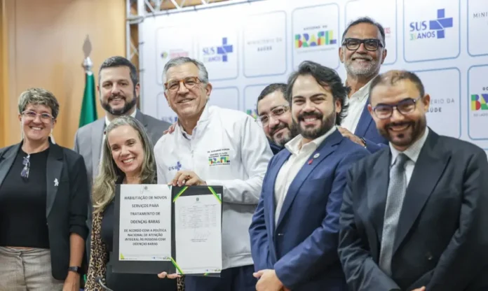 SUS terá exame de DNA que reduz diagnóstico para seis meses Uma foto de grupo com nove pessoas em um evento oficial. No centro, o Ministro da Saúde, Dr. Alexandre Padilha, e uma mulher seguram e exibem um documento oficial em uma pasta preta. O documento possui o título 'HABILITAÇÃO DE NOVOS SERVIÇOS PARA TRATAMENTO DE DOENÇAS RARAS' e menciona a 'POLÍTICA NACIONAL DE ATENÇÃO INTEGRAL ÀS PESSOAS COM DOENÇAS RARAS', com logomarcas do SUS e do Governo Federal. O Dr. Padilha veste um casaco branco com seu nome e cargo bordados. As outras sete pessoas, homens e mulheres, estão ao redor, sorrindo e olhando para a câmera. O fundo é um painel modular com repetições dos logos 'SUS 30 ANOS', 'MINISTÉRIO DA SAÚDE' e 'GOVERNO FEDERAL BRASIL', ao lado de uma bandeira do Brasil à esquerda.