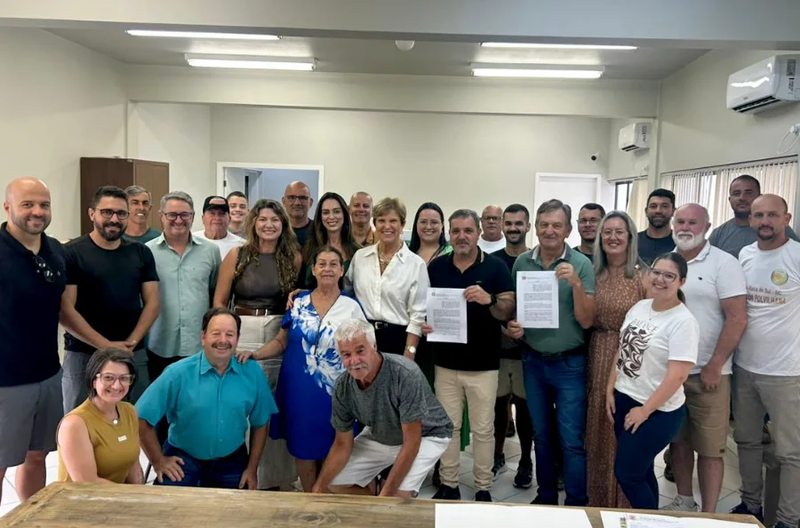 Prefeito de Santa Rosa do Sul, Almides Roberg da Rosa, e a Secretária de Estado da Assistência Social, Adeliana Dal Pont, seguram os convênios assinados para a construção de casas populares e uma residência de acolhimento, cercados por um grupo de autoridades locais e membros da comunidade no auditório municipal. A imagem marca a liberação de R$ 2,4 milhões em recursos estaduais para o município.