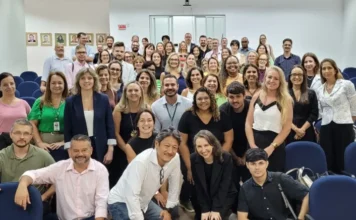 Saúde de SC debate inteligência artificial na gestão Fotografia em plano geral mostrando um grupo de aproximadamente 50 pessoas, homens e mulheres de diversas idades, posando sorridentes para uma foto coletiva dentro de um auditório. Eles são os profissionais e gestores da saúde pública de Santa Catarina participantes do primeiro evento sobre a transformação digital do SUS promovido pelo CIEGES. O grupo está organizado em várias fileiras, alguns sentados nas cadeiras azuis do auditório, outros em pé atrás e alguns agachados na frente. A sala tem paredes brancas e ao fundo uma porta de saída com uma placa vermelha "SAÍDA".