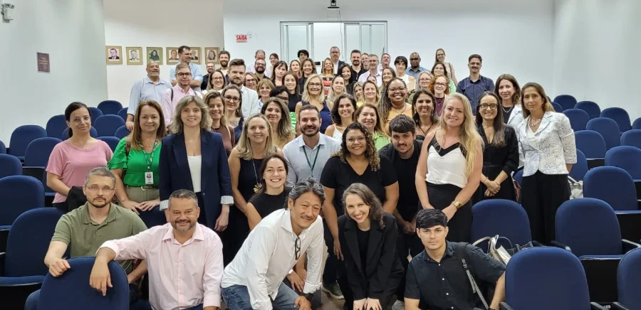 Fotografia em plano geral mostrando um grupo de aproximadamente 50 pessoas, homens e mulheres de diversas idades, posando sorridentes para uma foto coletiva dentro de um auditório. Eles são os profissionais e gestores da saúde pública de Santa Catarina participantes do primeiro evento sobre a transformação digital do SUS promovido pelo CIEGES. O grupo está organizado em várias fileiras, alguns sentados nas cadeiras azuis do auditório, outros em pé atrás e alguns agachados na frente. A sala tem paredes brancas e ao fundo uma porta de saída com uma placa vermelha 