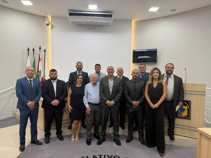 Scaini confirma renúncia e balanço de R$ 100 milhões Fotografia oficial capturada no plenário da Câmara de Vereadores de Balneário Arroio do Silva. Ao centro, o prefeito Evandro Scaini, vestindo terno cinza, está posicionado entre os vereadores e autoridades municipais. O grupo, composto por homens e mulheres em trajes formais, posa à frente da bancada diretiva, onde se veem as bandeiras oficiais e o brasão do Legislativo. O ambiente é iluminado e possui tons neutros, característico de sessões solenes.