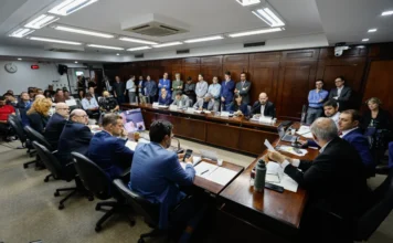 Secretário admite intenção de extinguir EGR em oitiva na CPI Vista geral da sala de reuniões durante a CPI das Concessões de Rodovias Estaduais na Assembleia Legislativa. Parlamentares e autoridades estão sentados ao redor de uma grande mesa de madeira em formato de U, ouvindo o depoimento do secretário Pedro Capeluppi (ao centro, ao fundo) sobre os custos de manutenção das estradas e a futura extinção da EGR. O público e a imprensa observam em pé ao fundo da sala.