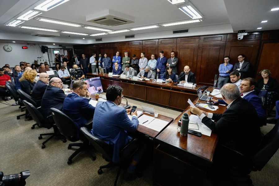 Vista geral da sala de reuniões durante a CPI das Concessões de Rodovias Estaduais na Assembleia Legislativa. Parlamentares e autoridades estão sentados ao redor de uma grande mesa de madeira em formato de U, ouvindo o depoimento do secretário Pedro Capeluppi (ao centro, ao fundo) sobre os custos de manutenção das estradas e a futura extinção da EGR. O público e a imprensa observam em pé ao fundo da sala.