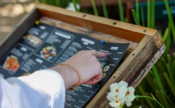 Setor de serviços de SC cresce 3,2% e supera média do país Uma mão feminina com pulseiras douradas aponta para opções em um cardápio de restaurante fixado em um suporte de madeira ao ar livre, exemplificando o consumo no setor de serviços prestados às famílias em Santa Catarina.