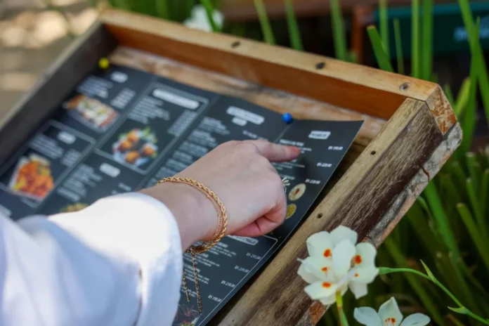 Setor de serviços de SC cresce 3,2% e supera média do país Uma mão feminina com pulseiras douradas aponta para opções em um cardápio de restaurante fixado em um suporte de madeira ao ar livre, exemplificando o consumo no setor de serviços prestados às famílias em Santa Catarina.