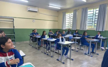 Siderópolis retoma aulas para 1,5 mil alunos da rede Fotografia de uma sala de aula iluminada na rede municipal de Siderópolis. Alunos de diferentes idades, vestindo uniformes azuis, estão sentados em suas classes organizadas em fileiras, prestando atenção ao início das atividades do ano letivo. Em primeiro plano, um estudante observa o ambiente, enquanto ao fundo outros colegas aguardam as instruções dos professores.