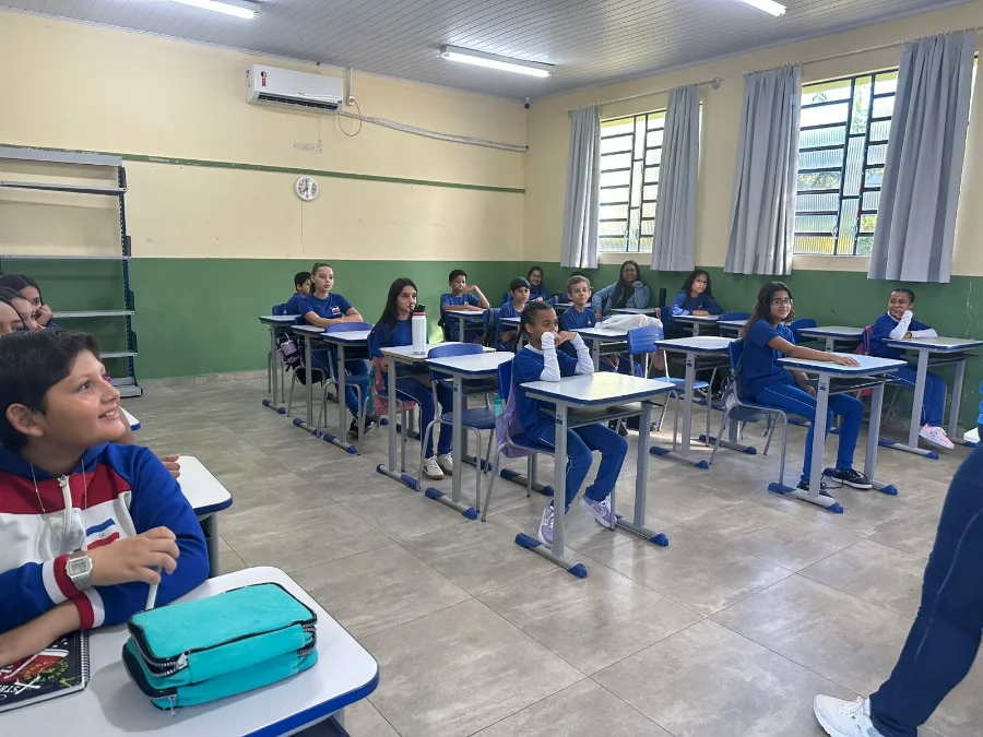 Fotografia de uma sala de aula iluminada na rede municipal de Siderópolis. Alunos de diferentes idades, vestindo uniformes azuis, estão sentados em suas classes organizadas em fileiras, prestando atenção ao início das atividades do ano letivo. Em primeiro plano, um estudante observa o ambiente, enquanto ao fundo outros colegas aguardam as instruções dos professores.