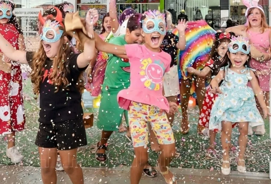 Grupo de crianças sorridentes celebrando o Carnaval no Sombrio Moda Shopping. Elas usam máscaras coloridas e fantasias enquanto se divertem em meio a uma chuva de confetes, ilustrando o clima festivo e de convivência do evento Folia Kids.