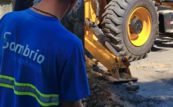 Sombrio Saneamento amplia rede de esgoto no bairro Raizeira Um colaborador da concessionária Sombrio Saneamento, vestindo uniforme azul com o logotipo da empresa, observa o trabalho de uma escavadeira em uma via urbana durante a instalação da rede de esgoto e pavimentação no bairro Raizeira.