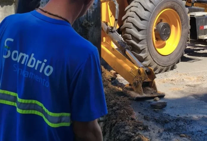 Sombrio Saneamento amplia rede de esgoto no bairro Raizeira Um colaborador da concessionária Sombrio Saneamento, vestindo uniforme azul com o logotipo da empresa, observa o trabalho de uma escavadeira em uma via urbana durante a instalação da rede de esgoto e pavimentação no bairro Raizeira.