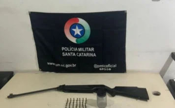 Suspeito é detido em Sombrio ao tentar vender espingarda Espingarda modificada e munições calibre .22 apreendidas pela Polícia Militar de Santa Catarina em Sombrio, dispostas sobre uma mesa com a bandeira da corporação ao fundo.