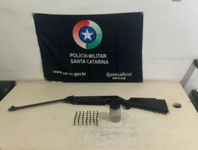 Suspeito é detido em Sombrio ao tentar vender espingarda Espingarda modificada e munições calibre .22 apreendidas pela Polícia Militar de Santa Catarina em Sombrio, dispostas sobre uma mesa com a bandeira da corporação ao fundo.